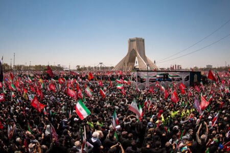 A wide-angle aerial shot capturing a massive crowd gathered in a large square in Tehran, featuring the Azadi Tower in the background and a sea of Iranian flags being held by attendees, reflecting the complex political atmosphere following the US-Israeli war against Iran.
