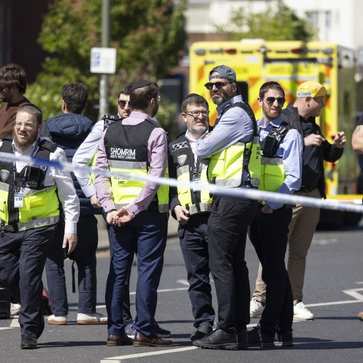 A Ceasefire Won’t End Iran’s Terror War on Europe A group of security personnel wearing high-visibility vests—some labeled with Shomrim North West London Emergency Response Unit—standing behind a white cordon tape on a city street with an ambulance in the background, showing emergency operations related to Iran's Terror War.