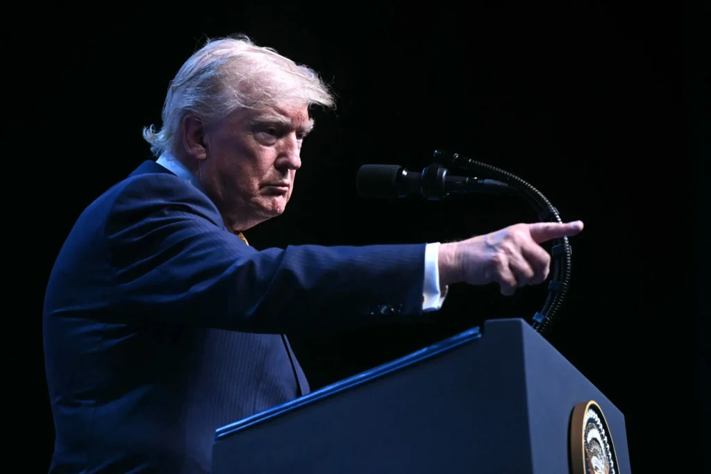 A side-angle shot of Donald Trump standing behind a dark podium with a microphone, pointing his index finger forward with a serious expression against a plain black background, illustrating a political stance to get tough with Iran.