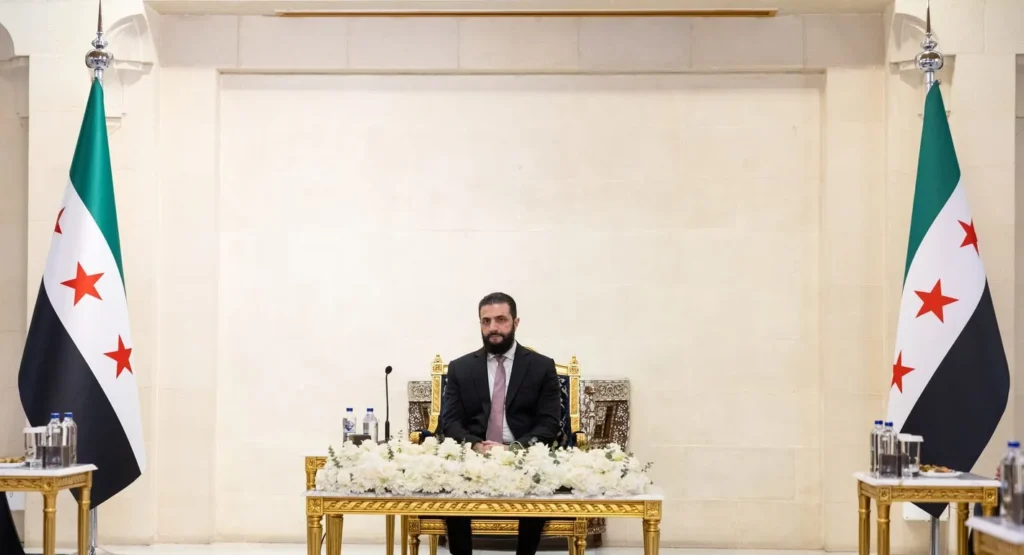 A New Patrimonialism is Undermining Syria’s Transition Syrian interim president Ahmed al-Sharaa in a dark suit sits at a low ornate table adorned with white flowers, flanked by two national flags of Syria against a plain indoor wall, framing political developments regarding Syria's Transition.