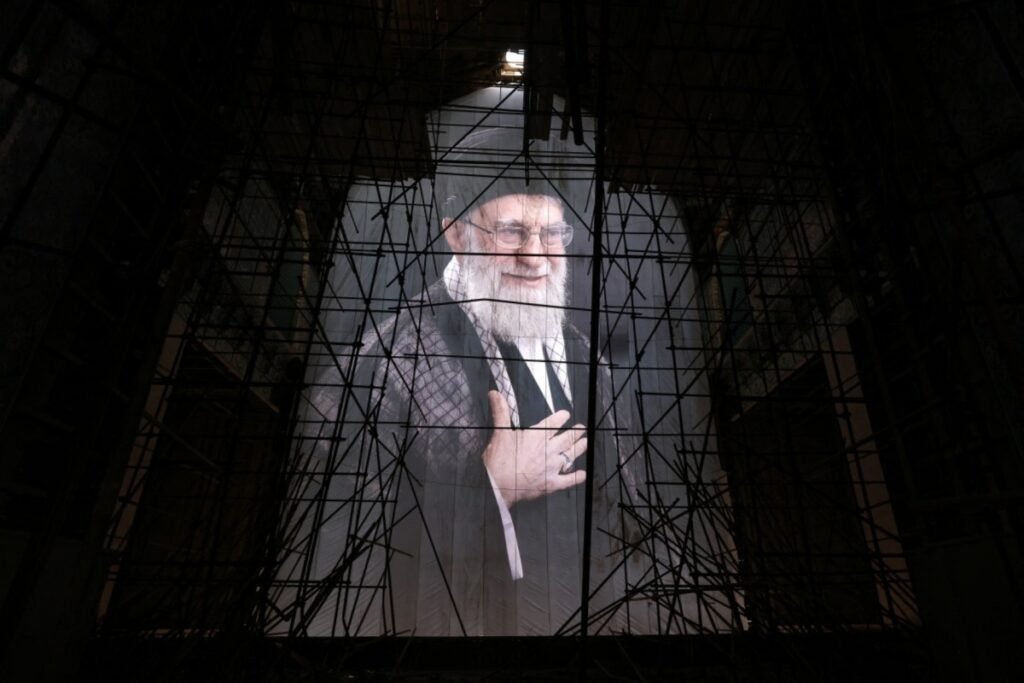 After Khamenei: the security state he built, and the son who inherits it A large portrait of Ali Khamenei smiling with his hand on his chest is visible through a complex network of dark scaffolding inside a dimly lit structure, symbolizing transitions After Khamenei.