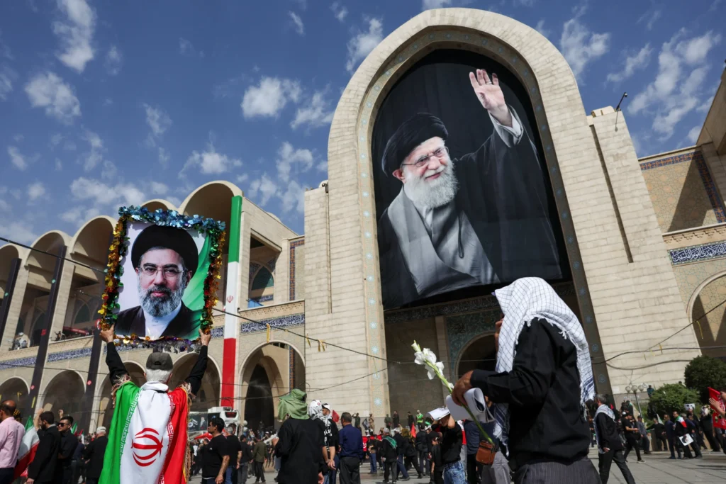 A wide outdoor shot in front of the Imam Khomeini Grand Mosalla building showing crowds of mourners, with one man draped in an Iranian flag holding aloft a framed portrait of Mojtaba Khamenei, while a massive banner of the late Ayatollah Ali Khamenei hangs over the main archway under a blue sky, capturing political transitions related to Trump's Iran Deal.