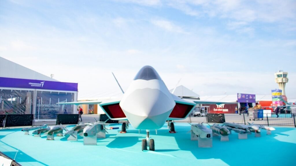Why NATO Depends on Turkey A head-on view of Turkey’s KAAN fifth-generation fighter jet displayed outdoors on a turquoise surface with assorted military missiles placed in front of it under a bright blue sky, underscoring the defense industrial base relevant to why NATO Depends on Turkey.