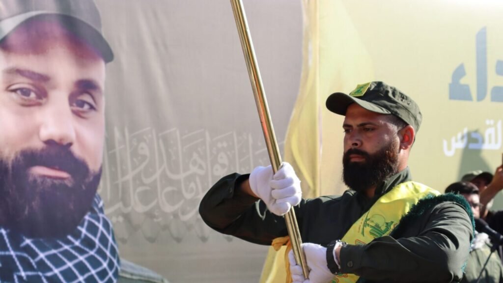 Turkey Hezbollah Alliance Deepens Amid the 2026 Iran War Now A Hezbollah member in uniform and white gloves holds a flag staff during a ceremony in front of a memorial portrait, highlighting the organizational presence linked to the evolving Turkey Hezbollah Alliance.