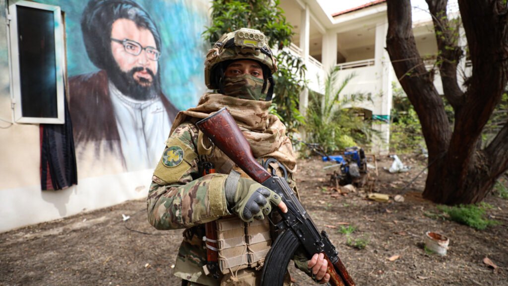 A masked soldier in tactical camouflage gear holding an assault rifle while standing in front of a wall mural depicting Hassan Nasrallah, highlighting regional tensions tied to Iran Syria Strikes.