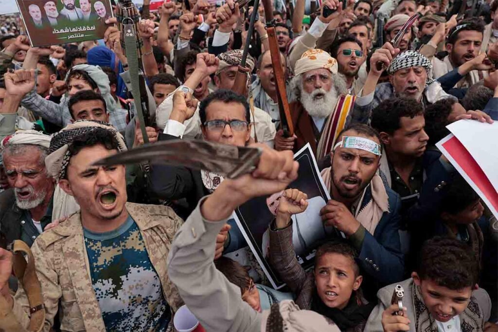 A dense crowd of Houthi supporters shouting and brandishing traditional daggers and rifles during a public rally, illustrating political rhetoric behind Houthis' threats.
