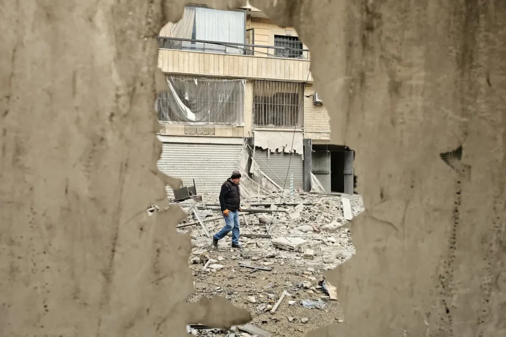 A man walking through rubble in front of a damaged building framing a hole in a broken wall, capturing post-conflict destruction and challenges in Rebuilding the Levant.