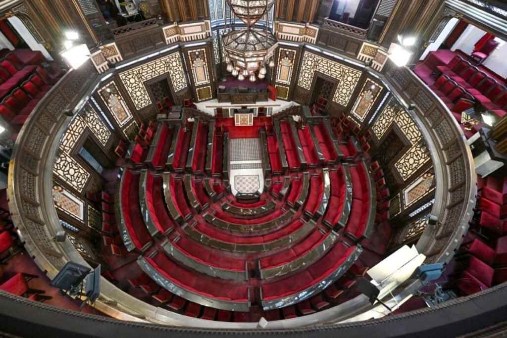 Syria’s parliament must now earn its legitimacy A high-angle, wide view looking down at the semi-circular assembly hall of a parliament building with rows of red velvet seats and intricate geometric wall patterns beneath a large chandelier, capturing the formal setting of Syria's parliament.