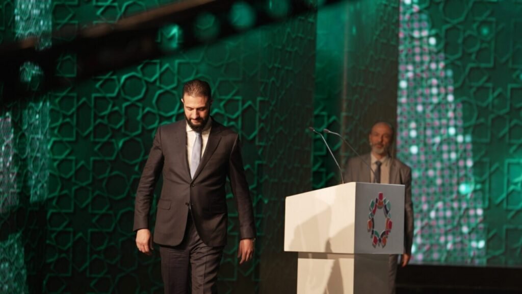 أحمد الشراعا يحتاج إلى تصحيح المسار Syrian leader Ahmed Al-Sharaa wearing a dark suit and light blue tie walks across a stage with his head cast downward, away from a white podium, against a backdrop of green illuminated geometric patterns, reflecting the political pressure demanding an immediate course correction.