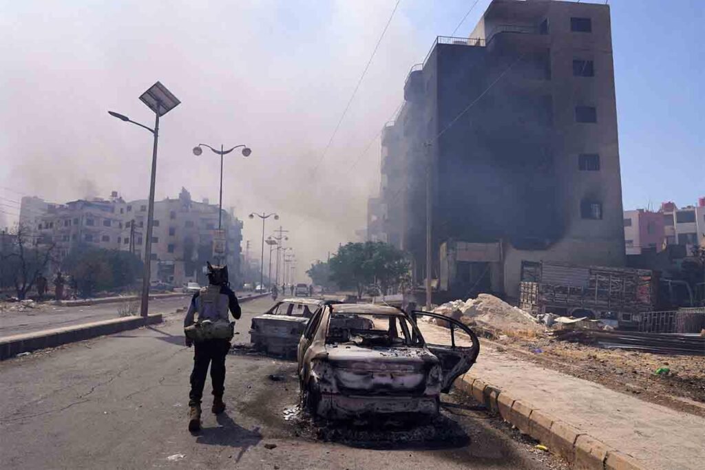One year after the massacres: Druze in Syria remain under acute threat An armed soldier walks past burnt and destroyed cars on a debris-littered street next to a charred apartment building emitting smoke, depicting the aftermath of conflict affecting the Druze in Syria.