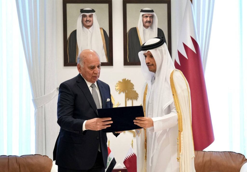 The Qatar Model: The Future of Iraq-GCC Economic Integration? An indoor official meeting showing a man in a business suit holding a document folder alongside a man wearing traditional Qatari attire and bisht, with official portraits and the flag of Qatar visible in the background, highlighting diplomatic steps toward Iraq-GCC.