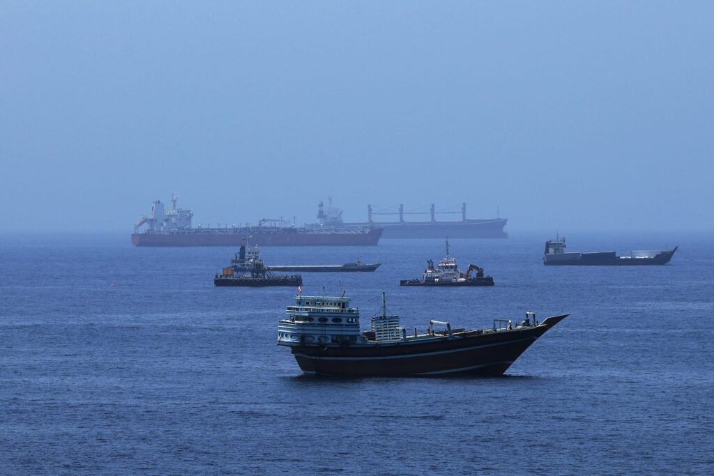 A traditional wooden dhow boat floats in the foreground with multiple commercial cargo ships and large oil tankers anchored on the calm blue sea under a clear sky, illustrating maritime activity heading toward the Hormuz Strait.
