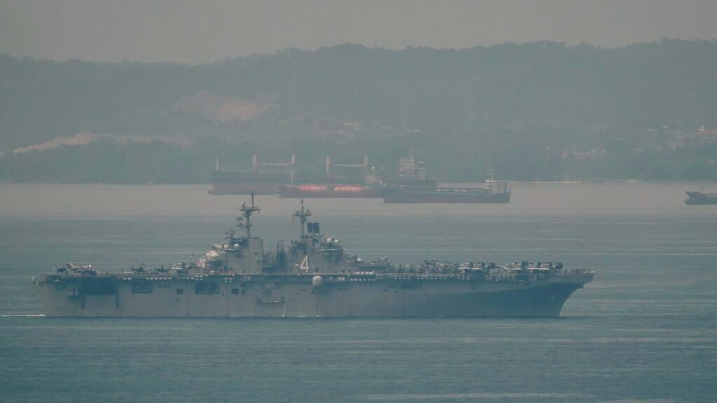 The amphibious assault ship USS Boxer navigating hazy coastal waters with cargo vessels in the background, illustrating operational strain tied to US Navy Overstretch.