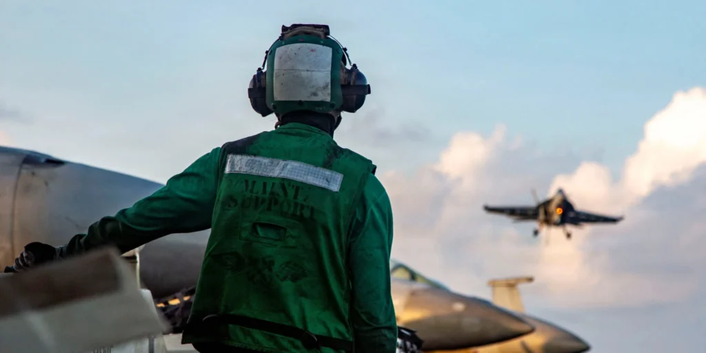 A military flight deck crew member wearing a green maintenance support vest and helmet watches from behind as a fighter jet prepares to land on an aircraft carrier against a cloudy sky, framing regional deterrence strategies regarding Iran.