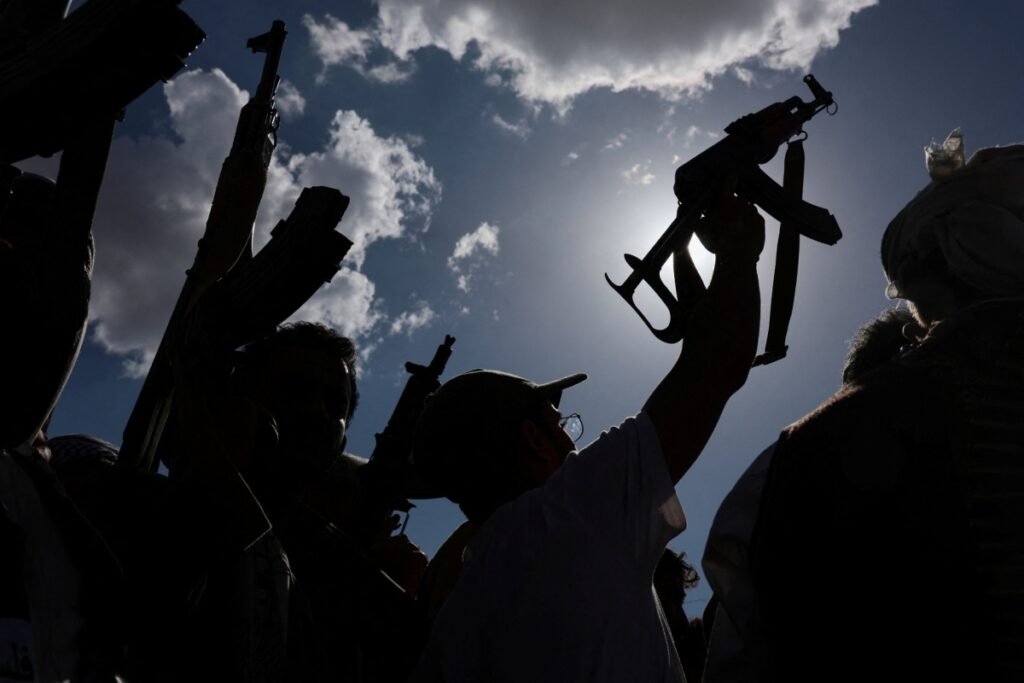 A dramatic low-angle silhouette shot of individuals at a rally, with one person in the foreground raising an assault rifle against a bright, cloudy sky, illustrating the intensified Fighting in Yemen linked to broader regional conflicts.