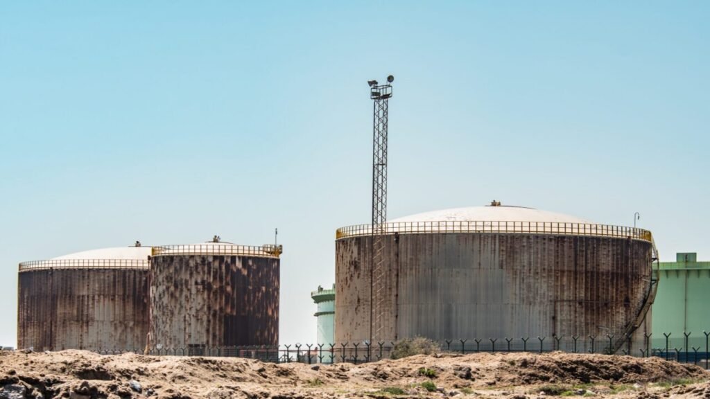 Large industrial oil storage tanks and facility infrastructure behind a security perimeter fence, representing energy vulnerabilities during discussions on Iran Bombing Kuwait.