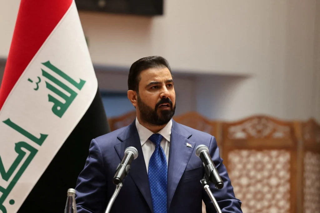 نخستوزیر جدید عراق باید عراق را به واشنگتن بدبین بفروشد A medium shot of Iraq's new prime minister, Ali al-Zaidi, wearing a dark blue suit and a patterned blue tie while speaking at a podium with two microphones, with the Iraqi national flag partially visible on the left, representing Iraq's new prime minister.