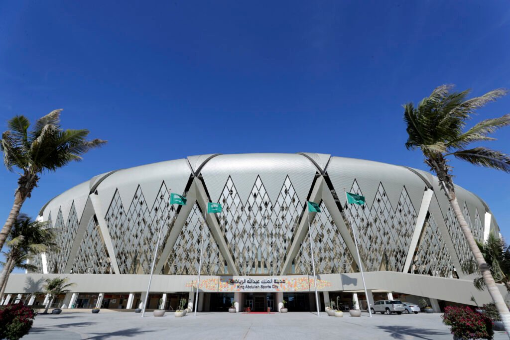 A Saudi Plus World Cup 2034: A Chance for Gulf Unity and Resilience Exterior view of the modern King Abdullah Sports City stadium architecture in Jeddah featuring palm trees and Saudi flags under a clear blue sky, highlighting sports infrastructure for World Cup 2034.
