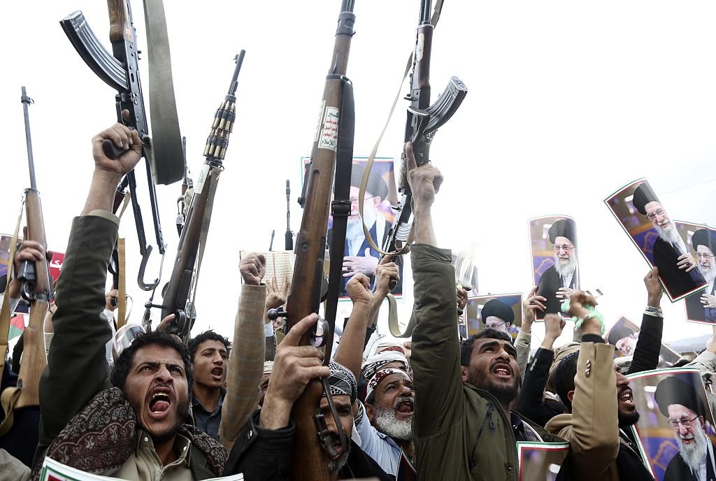 How the Houthis Could Put the World in Double Dire Straits A crowd of men chanting passionately while lifting assault rifles and wooden-stocked firearms into the air, holding up portraits of a religious leader against a bright sky, illustrating the militant mobilization of the Houthis.