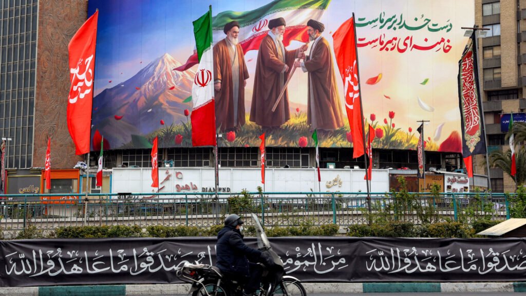 بين الردع والدبلوماسية: معضلة الخليج وإيران A street scene featuring a large urban mural displaying portraits of Ruhollah Khomeini, Ali Khamenei, and Mojtaba Khamenei alongside Iranian flags, setting a backdrop for regional security conversations on the Gulf Iran dilemma.
