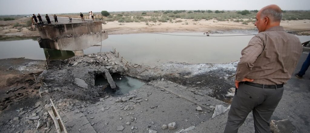 A man standing on the edge of a destroyed bridge overlooking rubble and a river, illustrating infrastructure damage caused by Iran conflict escalation.