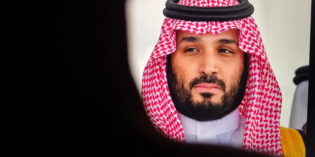 A close-up shot of Saudi Crown Prince Mohammed bin Salman wearing a traditional red and white checkered ghutra, looking thoughtfully to the side as framed through a dark foreground silhouette in 8.webp, reflecting the strategic considerations of a regional security guarantor.