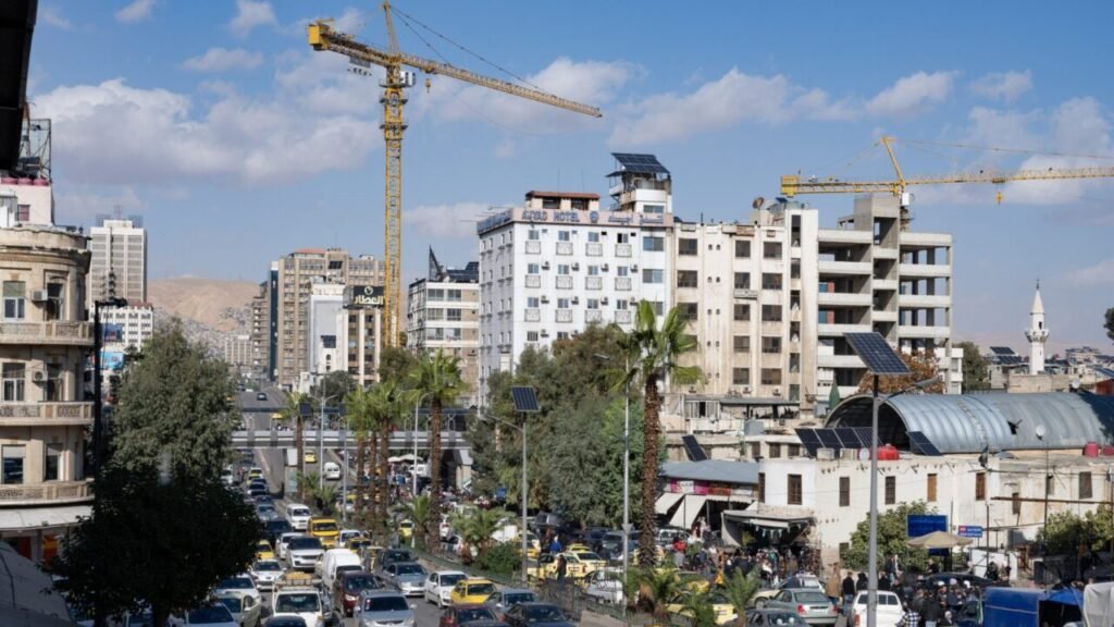 Heavy traffic moving along a busy city street in central Damascus with tall yellow construction cranes operating above modern high-rise buildings, symbolizing urban reconstruction efforts shaping Middle East's Future.