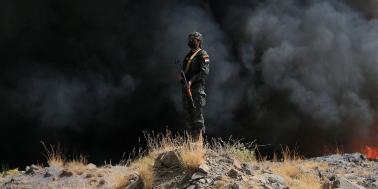 حصار الحوثيين للمملكة العربية السعودية يفتح جبهة جديدة لإيران An armed soldier holding a rifle standing guard on rocky ground against a dense backdrop of heavy black smoke and flames, highlighting regional tension regarding the Houthi Blockade of Saudi Arabia.