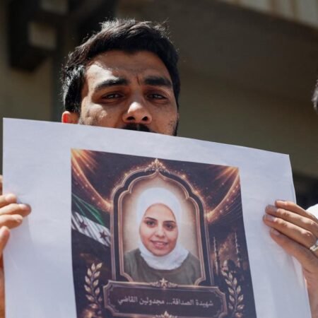 A close-up photograph of a man solemnly holding a framed memorial poster of a woman wearing a white hijab, with the Syrian flag emblem visible on the poster, symbolizing the public mourning and dissent that restrictive Syria Speech Laws threaten to silence, thereby hindering the fragile dialogue essential for Post-Assad reconstruction.