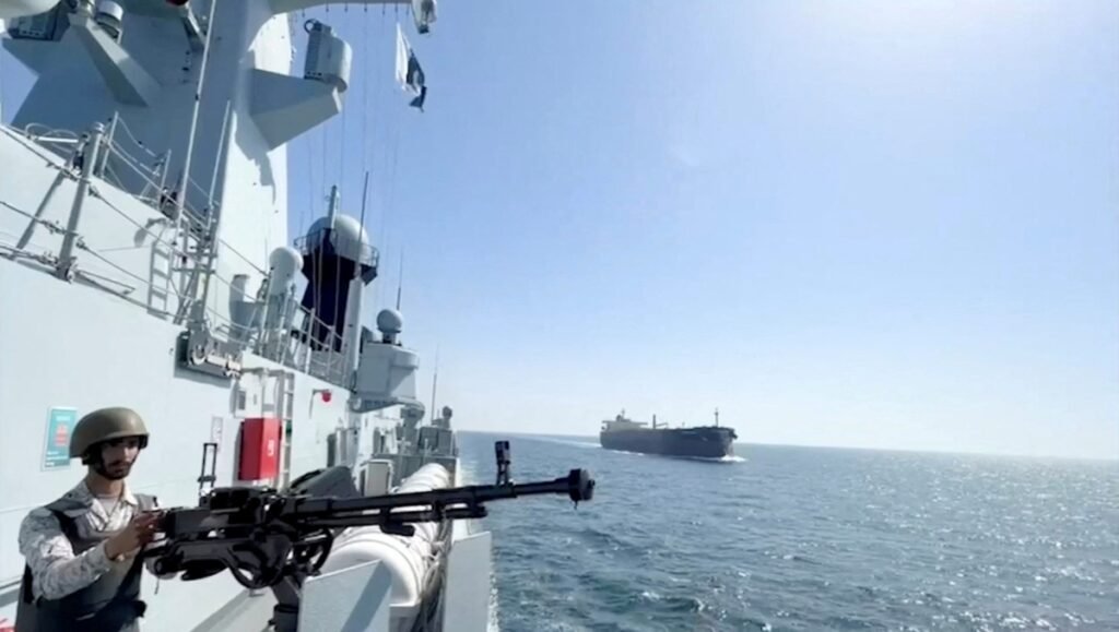 Pakistan Red Sea policy shifts toward Saudi alliance A Pakistani Navy sailor, wearing a helmet and tactical vest, is pictured manning a heavy machine gun aboard a warship, looking out over the sea towards a distant cargo vessel. This image visually contextualizes the recent geopolitical analysis surrounding the Pakistan Red Sea policy, as the navy asserts its presence in a region of heightened tension. The Pakistani flag is visible on the ship's mast.
