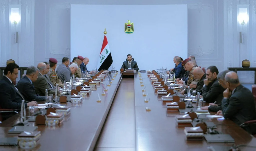 Iran influence in Iraq blocks al-Zaidi’s state control bid High-level Iraqi civilian and military officials gathered around a long conference table during a formal government security meeting in Baghdad, with the national flag and emblem prominently displayed, visually representing the internal governance challenges where Iran influence in Iraq continues to obstruct Prime Minister al-Zaidi's bid to establish full state control.