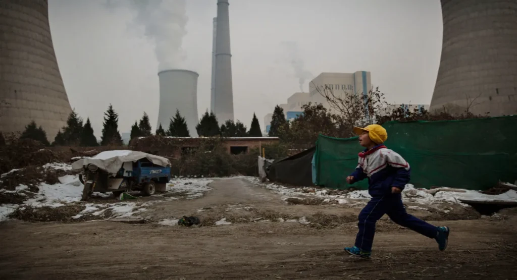 A child running along a dirt path near an industrial power plant with tall cooling towers emitting steam, reflecting the domestic industrial infrastructure driving China's Energy Security through electrification.