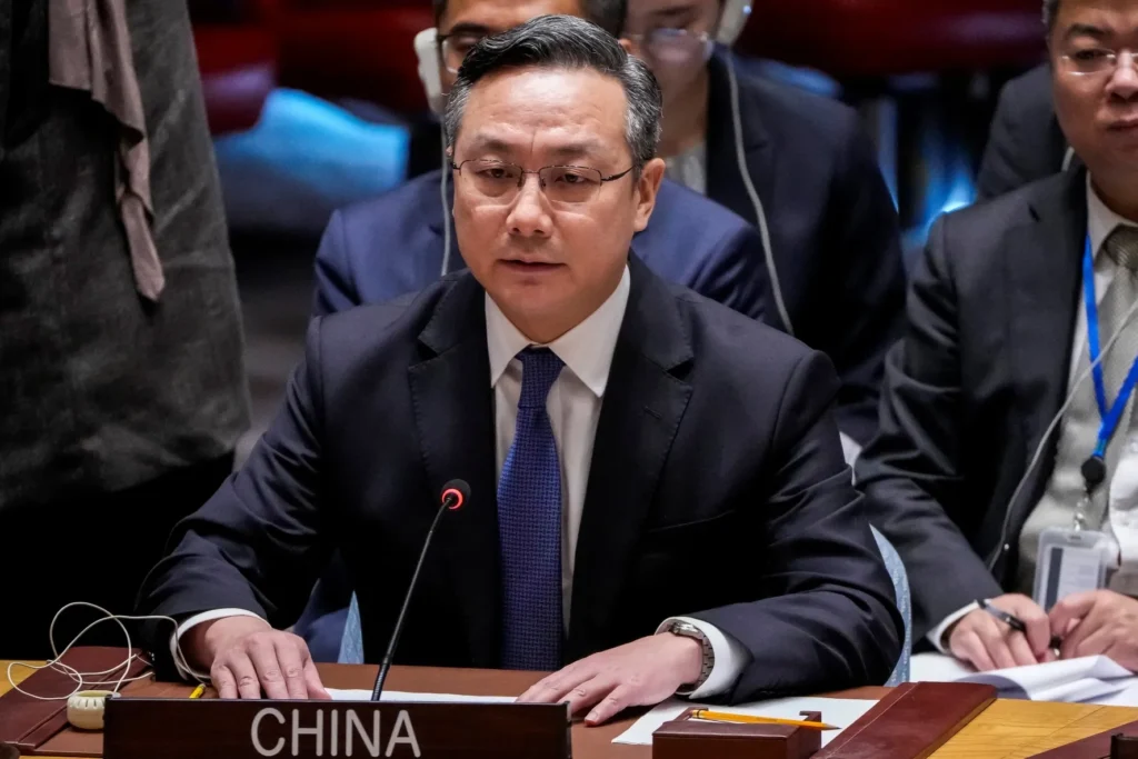 A close-up photograph of a senior Chinese diplomat wearing a dark suit, white shirt, and blue tie, speaking into a microphone at a UN Security Council session with a "CHINA" placard prominently displayed in the foreground, symbolizing the diplomatic moderation of the China Iran stance that effectively limits broader war risks to global trade routes and energy security.