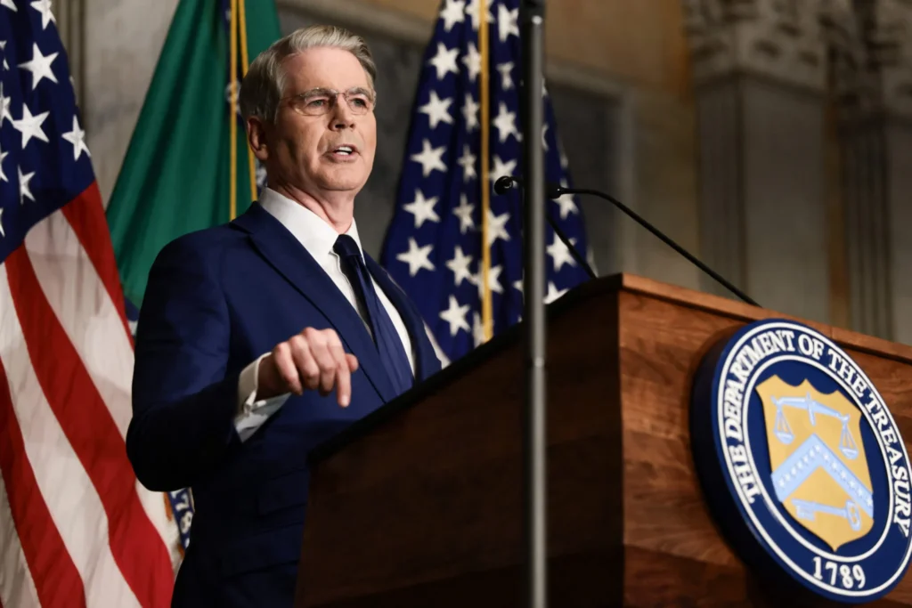 Operation Economic Outcast Targets Iran Lifelines A photograph of US Treasury Secretary Scott Bessent speaking emphatically at a wooden podium, pointing his finger forward with the official seal of the Department of the Treasury and US flags in the background, visually representing the financial pressure campaign of Operation Economic Outcast that aims to sever Iran's critical economic lifelines.