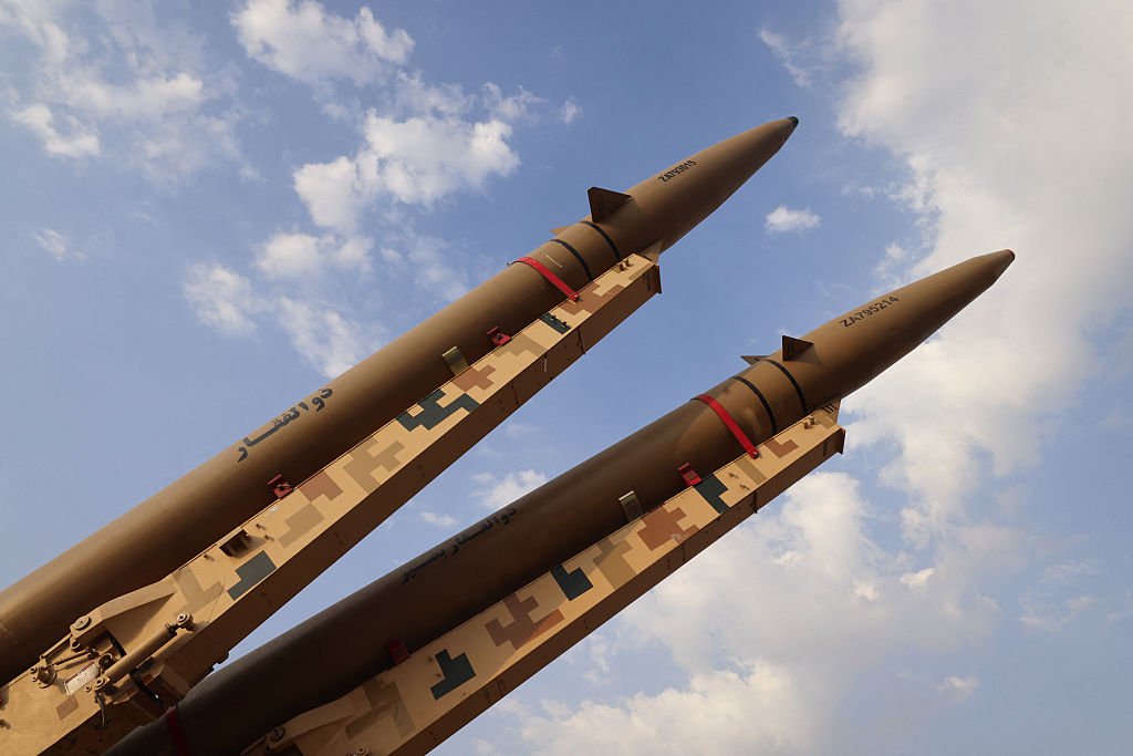 Iran Ballistic Missile Production Expands Underground A low-angle photograph showing two large, sand-colored ballistic missiles secured with red retaining bands on desert-camouflaged mobile launchers, angled sharply against a bright blue sky with scattered clouds, visually affirming the operational readiness and growing inventory resulting from Iran Ballistic Missile Production, which defense analysts report is increasingly being shifted and expanded into fortified underground facilities to protect it from potential preemptive strikes.