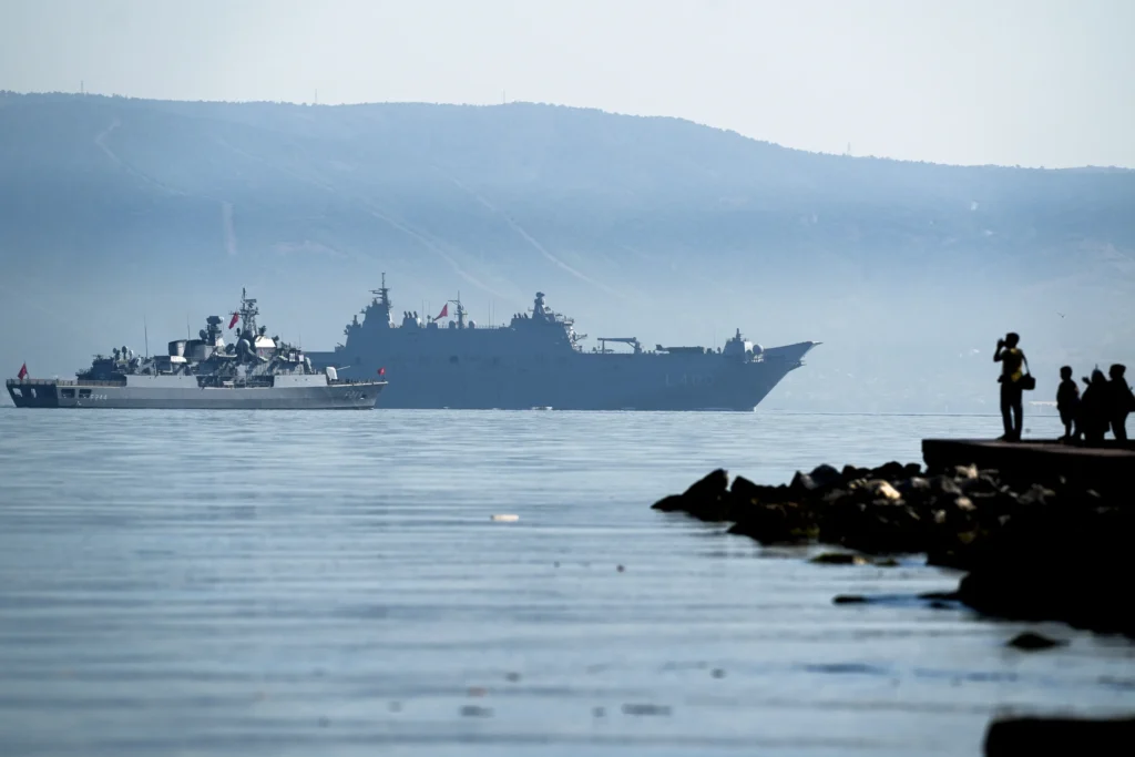 A wide maritime photograph showing two grey naval warships, including a large amphibious assault vessel, sailing through calm blue waters along a hazy, mountainous coastline, while the dark silhouettes of bystanders observe from a rocky pier, visually capturing the coastal presence and naval dynamics that are central to the evolving layered integration now shaping Middle East Security.