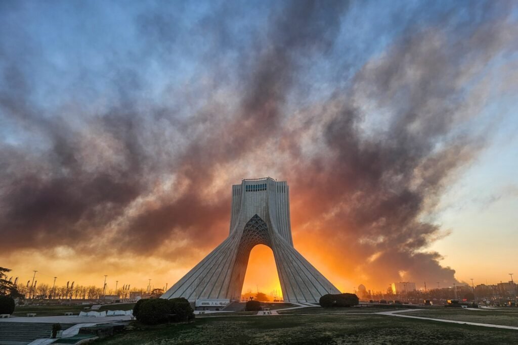 Dramatic photograph of the Azadi Tower in Tehran at dusk, with massive, dark plumes of smoke rising from the horizon behind the iconic structure. This volatile scene visually underscores the geopolitical analysis concerning Iran Postwar Strategy and regional security dynamics.