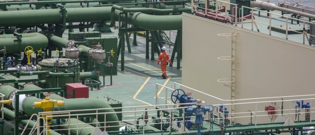 Pakistan LNG Crisis Deepens Amid Hormuz Strait Closure A high-angle photograph of an LNG carrier deck showing a worker in an orange safety suit walking amidst complex green pipelines, valves, and structural tanks, illustrating the fragile maritime infrastructure at the heart of the Pakistan LNG crisis as the closure of the Strait of Hormuz threatens the nation's vital energy supply chains.