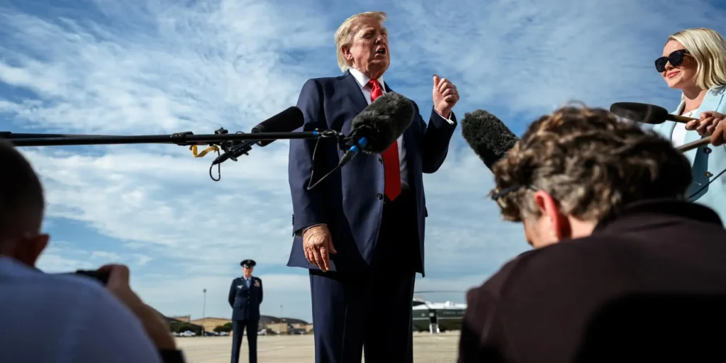 A low-angle photograph of US President Donald Trump speaking emphatically outdoors to a crowd of reporters, with prominent microphone booms in the foreground, while a woman in sunglasses and a US Air Force officer stand in the background under a cloudy blue sky, visually capturing the intense public pressure and political messaging surrounding the Trump Iran War Freeze and its potential backfire ahead of the midterms.