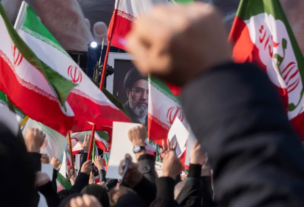 Iran Hardliners Retain Control in Government Reshuffle Demonstrators raise clenched fists and wave Iranian flags before a portrait of the Supreme Leader, reflecting how Iran hardliners maintain political dominance.