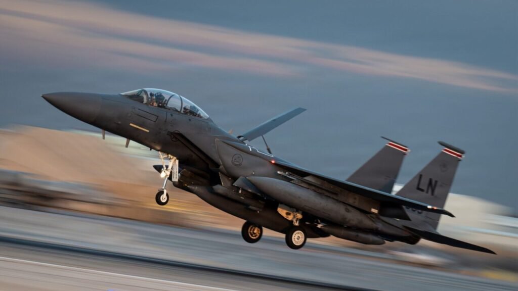 A dynamic panning photograph of a US Air Force F-15E Strike Eagle with its landing gear lowered, captured in motion blur against a twilight sky at a Middle Eastern airfield, symbolizing the rapid airpower projection and forward presence of US Military Bases in the Gulf that are essential for deterring Iranian aggression and securing Persian Gulf security.