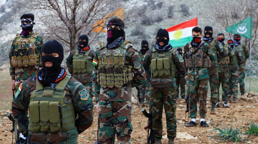 Kurdish Intervention in Iran Collapses Before It Begins A line of masked Kurdish fighters in woodland camouflage uniforms and heavy tactical chest rigs standing at attention on a rocky, arid hillside, with the Kurdish sun flag and a green militia flag displayed prominently in the background, symbolizing the paramilitary forces whose planned Kurdish intervention into Iranian territory collapsed politically and logistically before a single shot could be fired, underscoring the strategic failure detailed in the article.