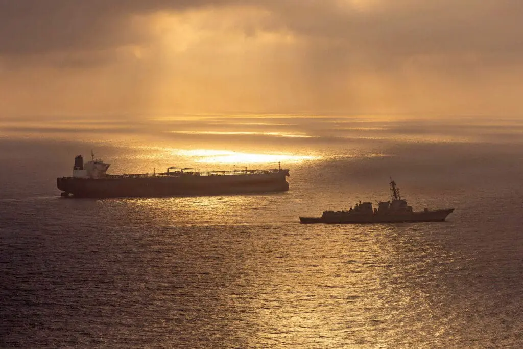 Wartime Oil Prices Defy High Forecasts Amid Conflict A serene golden sunset silhouettes a massive crude oil tanker being escorted by a naval warship across calm waters, symbolizing the military protection and geopolitical conflict risks that typically inflate Wartime Oil Prices, which are paradoxically defying high market forecasts amid the current tensions.