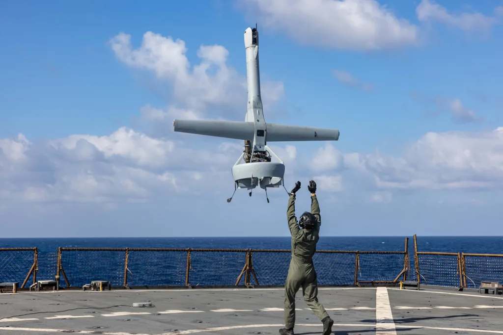 إعادة بناء الترسانة الأمريكية تتسارع لردع حرب الصين وتايوان A dramatic mid-air photograph showing a U.S. Marine in olive flight deck gear and helmet with raised arms, launching a grey V-BAT vertical takeoff and landing unmanned drone from the flight deck of the USS Carter Hall in the Mediterranean Sea, symbolizing the rapid deployment of next-generation tactical naval technology that defines the urgency of the American Arsenal Rebuild as military planners race to bolster expeditionary deterrence against a potential China-Taiwan war.