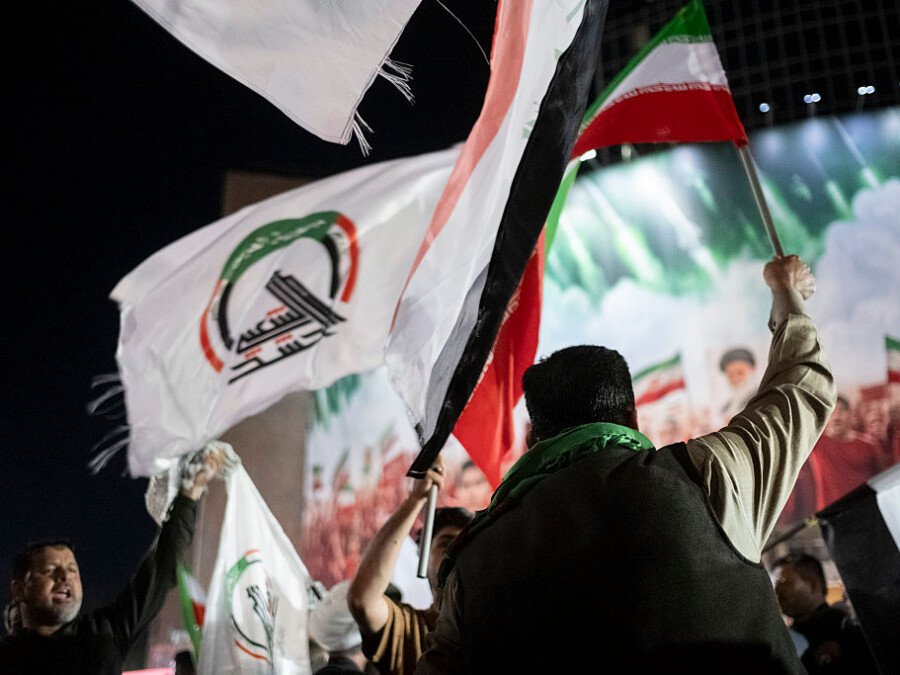 الميليشيات العراقية تتحدى نزع السلاح بينما تحمي طهران الأسلحة A nighttime photograph of a crowded political rally where a man raises an Iraqi national flag alongside a white militia flag with a green emblem, while an Iranian flag waves in the background, visually representing the unified defiance of Iraq militias against disarmament initiatives as Tehran continues to shield their weapons supply.