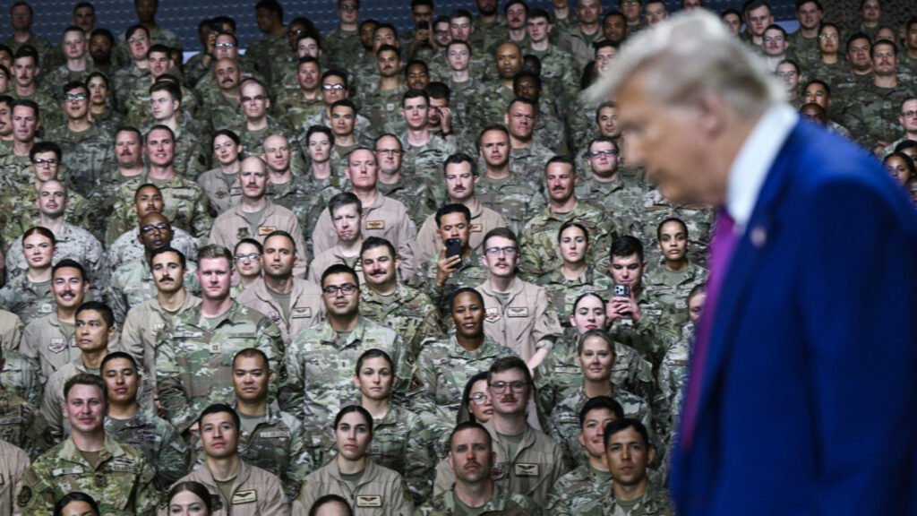 تحول القواعد الأمريكية في الخليج من دروع إلى أهداف في الحرب ضد إيران A photograph showing President Donald Trump as a blurred figure in the foreground, facing a dense crowd of US military personnel in camouflage uniforms, visually representing the strategic shift of US Gulf Bases from protective shields into primary targets for Iranian retaliation in a broader conflict.