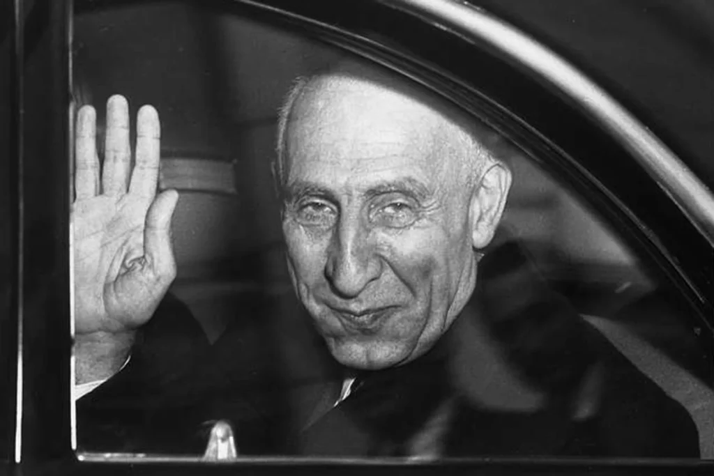 1953 Iran Coup Still Fuels Deep US-Iran Hostility A historical black-and-white photograph of former Iranian Prime Minister Mohammad Mosaddegh solemnly waving from a car window, symbolizing the deep historical trauma of the 1953 Iran Coup that continues to fuel profound and enduring US-Iran hostility today.
