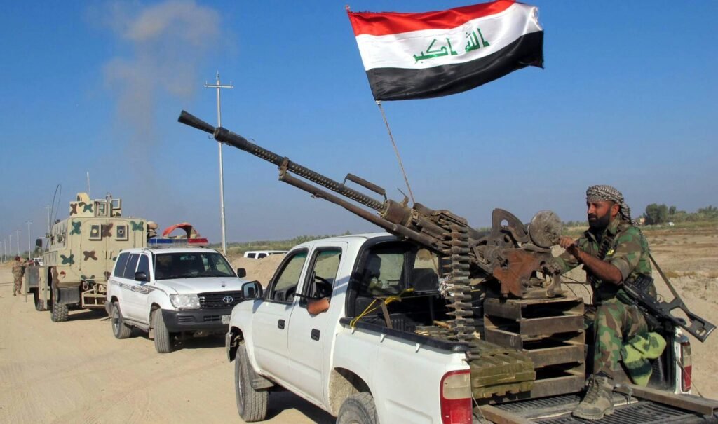 كيف يمكن منع الميليشيات من تعطيل العلاقات العراقية السعودية An armed fighter manning a heavy machine gun on a pickup truck flying the Iraqi flag in a military convoy, highlighting security threats that risk disrupting Iraqi-Saudi Relations.