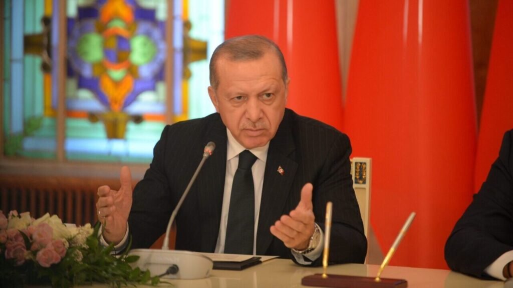 A close-up photograph of Turkish President Recep Tayyip Erdoğan speaking emphatically at a podium, gesturing assertively with both hands while wearing a dark suit and tie, set against an official backdrop of the red Turkish flag and stained glass, visually capturing the authoritative and unyielding political posture reflecting how the Kurdish Gambit strategically fuels his autocratic power grab and consolidation of executive authority.