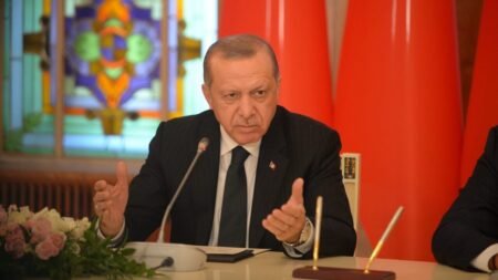 A close-up photograph of Turkish President Recep Tayyip Erdoğan speaking emphatically at a podium, gesturing assertively with both hands while wearing a dark suit and tie, set against an official backdrop of the red Turkish flag and stained glass, visually capturing the authoritative and unyielding political posture reflecting how the Kurdish Gambit strategically fuels his autocratic power grab and consolidation of executive authority.